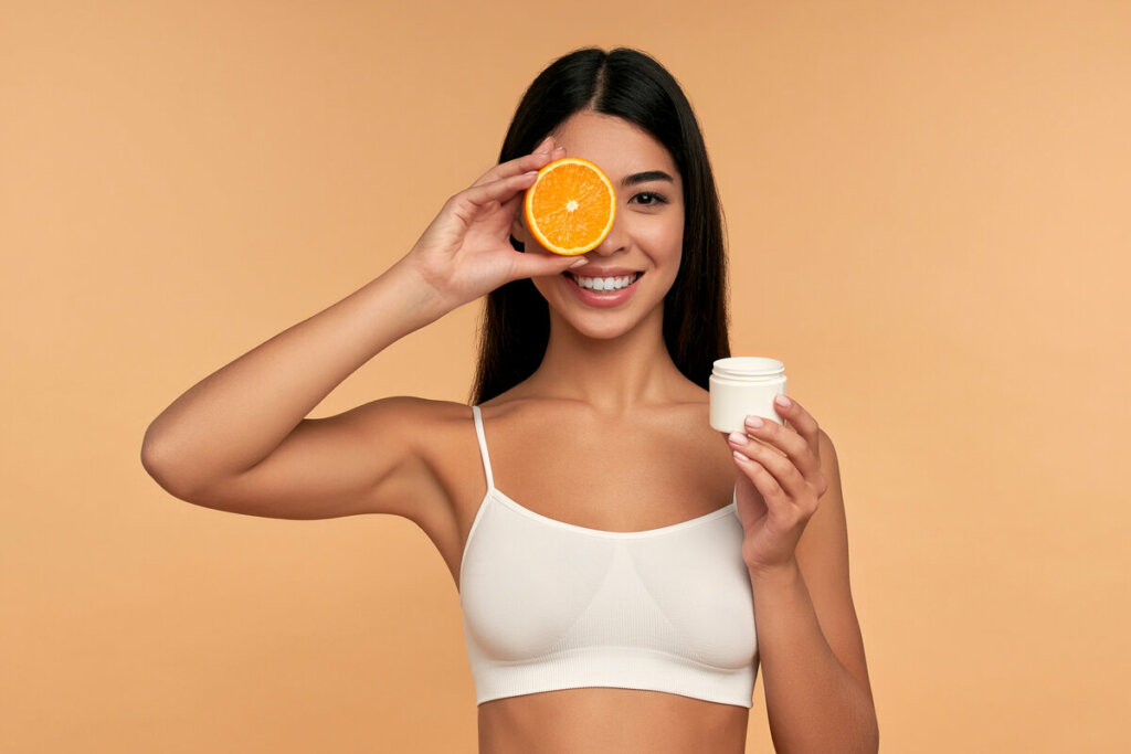 Mulher com top branco sorrindo segurando um recipiente branco e uma rodela de laranja em frente ao olho