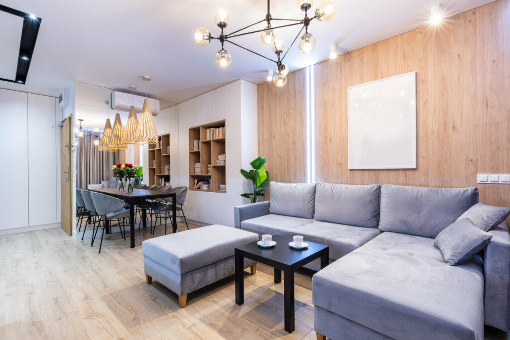 Sala de estar integrada com cozinha com sofá cinza, lustre e parede de madeira
