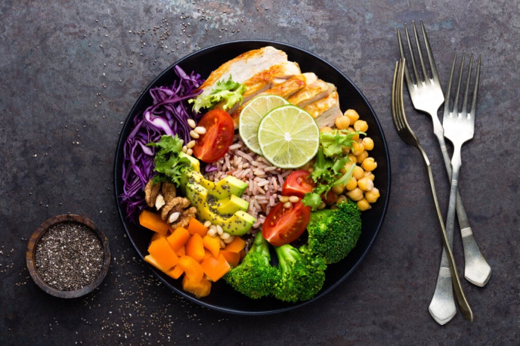 Prato com filé de frango, arroz integral, abacate, pimenta, tomate, brócolis, repolho vermelho, grão de bico, salada de alface fresca, pinhões e nozes.