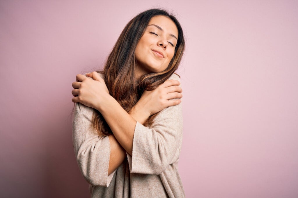 Mulher vestindo suéter casual em pé sobre fundo rosa abraçando-se feliz