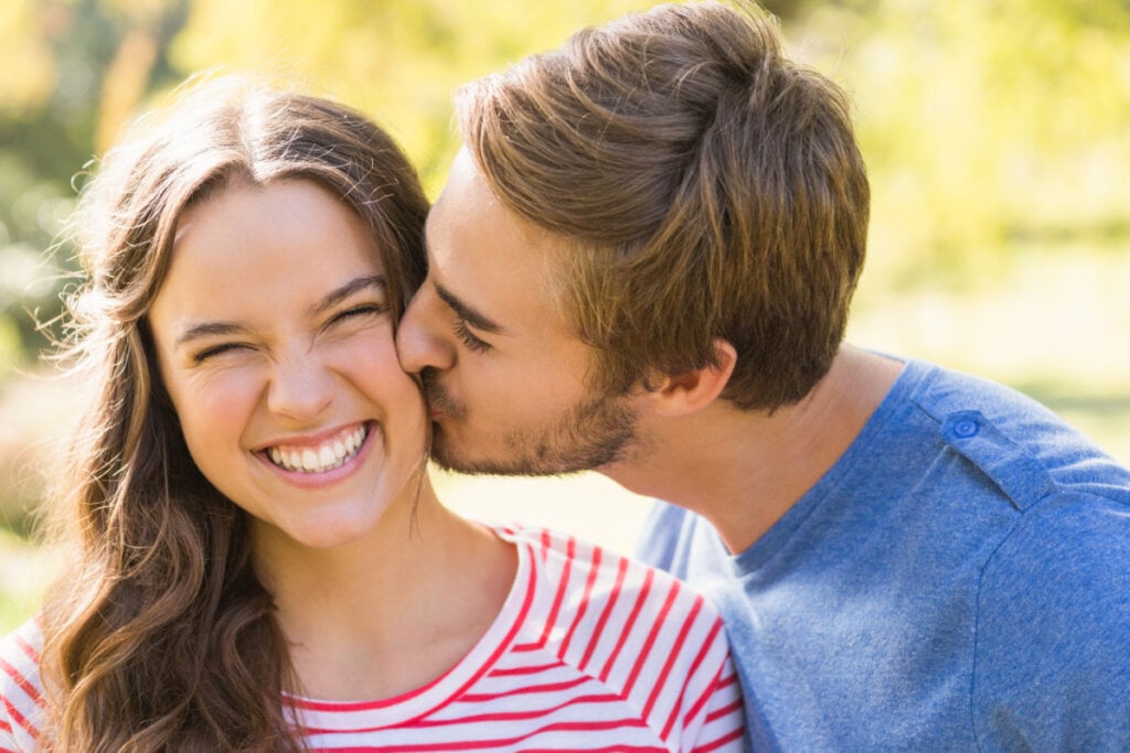 Homem dando beijo na bochecha de mulher sorridente em um parque.