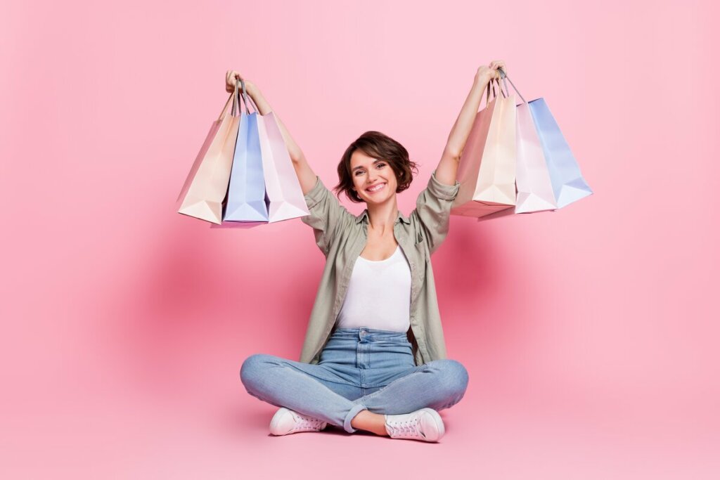 Mulher sentada segurando sacola de compras e sorrindo em frente a um fundo rosa