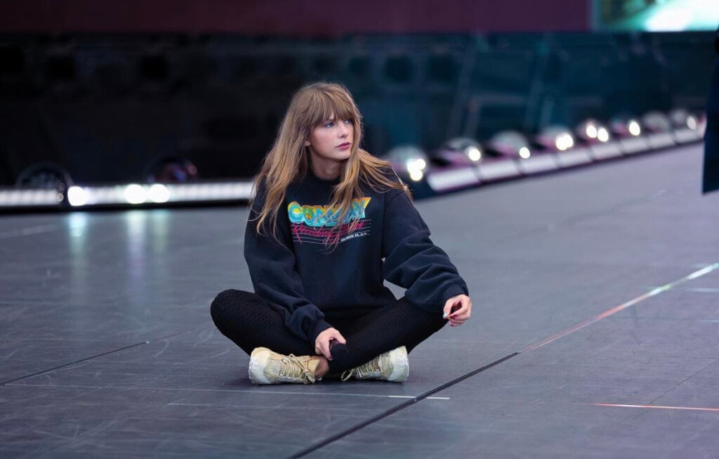 Taylor Swift de moletom sentada em estrutura de palco para preparação de turnê