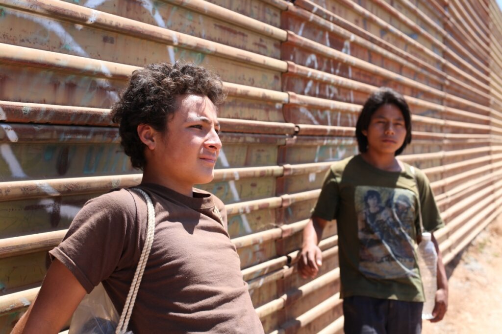 Cena do filme La Jaula de Oro, com dois jovens de semblante pensativo encostados em muro de ferro.