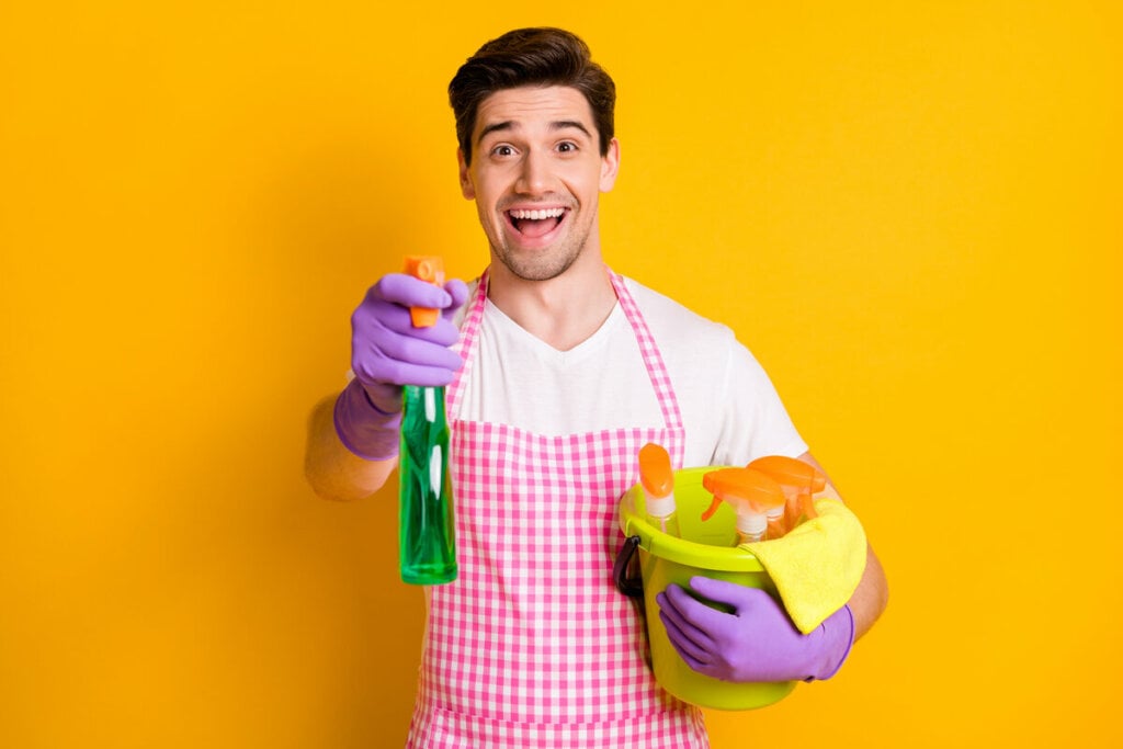 Homem com roupa de faxina e luva roxa sorrindo em um fundo amarelo