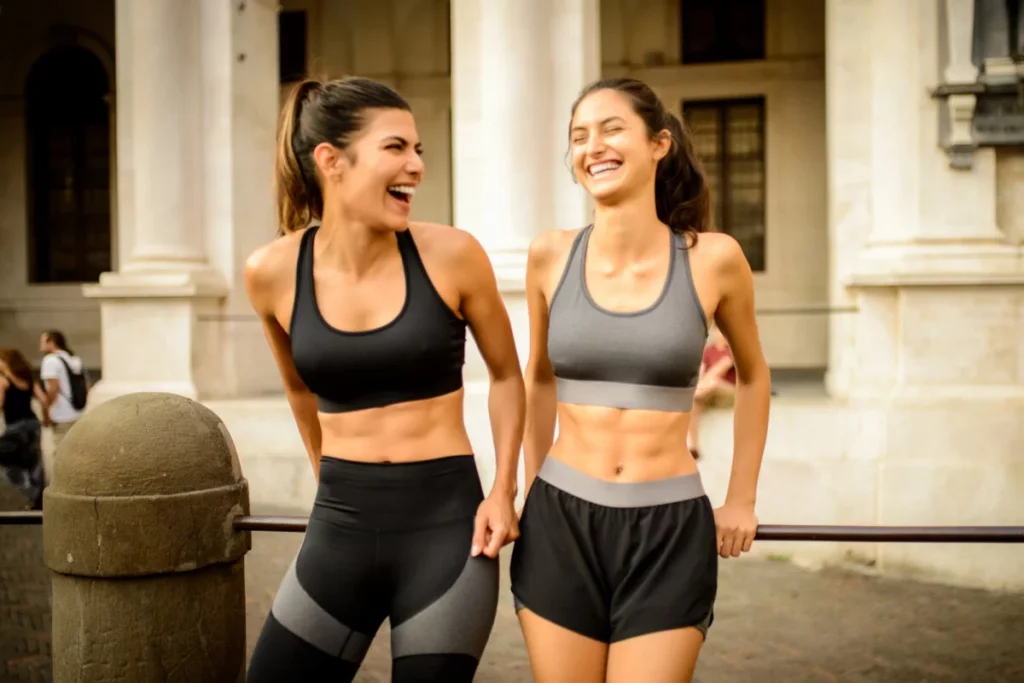 Duas mulheres sorridentes com roupas de academia. Ambas possuem barriga definida.