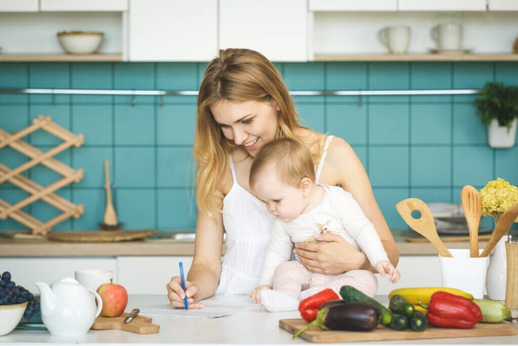 Mãe anotando algo em papel com bebê ao lado e frutas e vegetais em tábua próxima