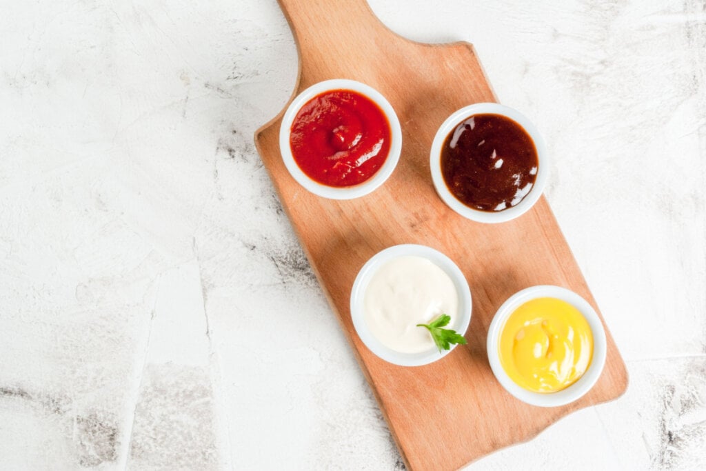 Conjunto de molhos em pires brancos: mostarda amarela, ketchup, molho barbecue e maionese. 