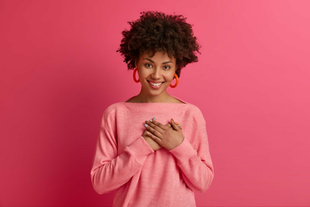 Mulher com blusa e brinco rosa com a mão no coração e sorrindo