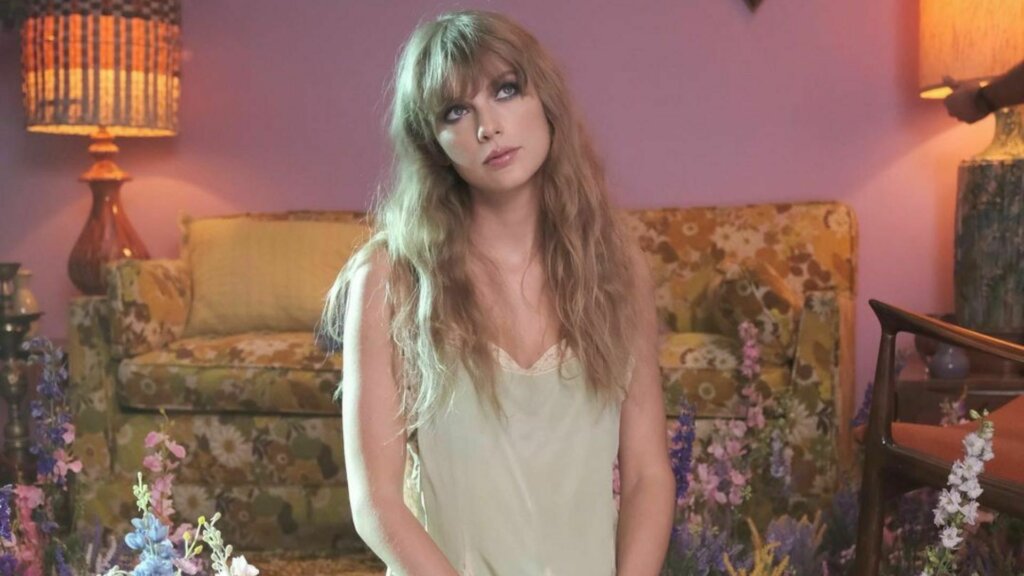 Imagem do clipe Lavander Haze, de Taylor Swift. A cantora veste uma blusa branca e está em cenário de sala de estar com parede lilás.