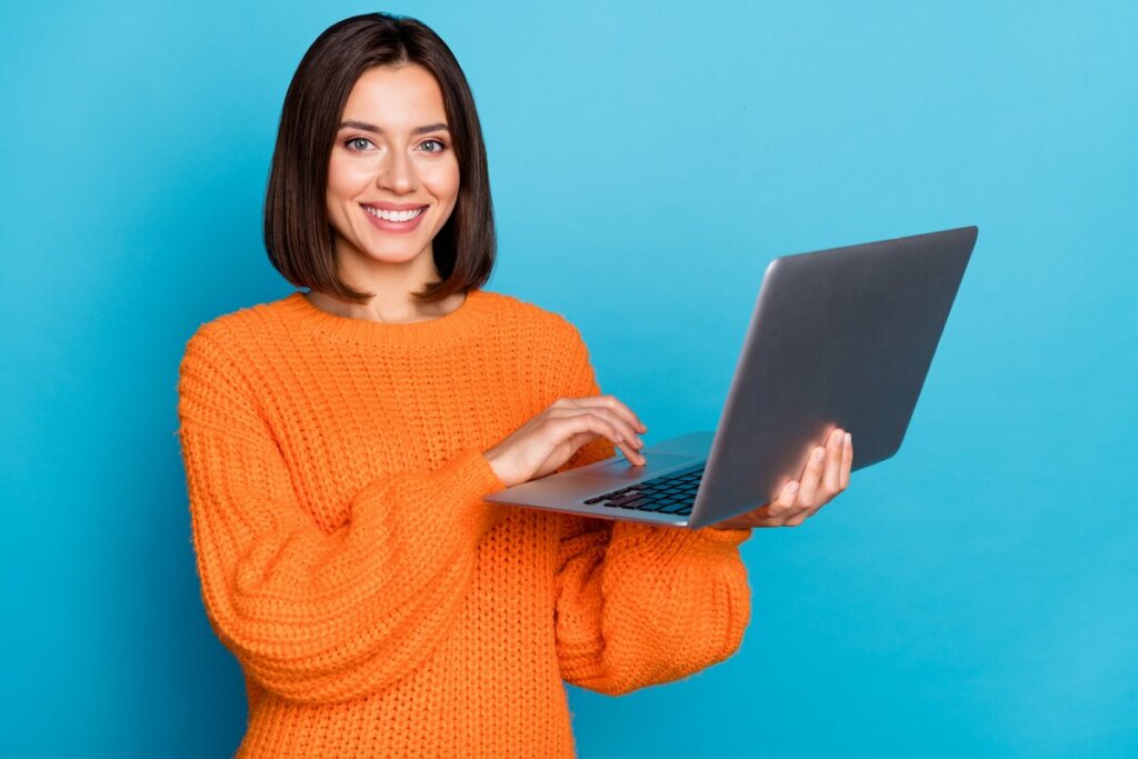 Mulher de blusa laranja sorrindo e segurando um notebook