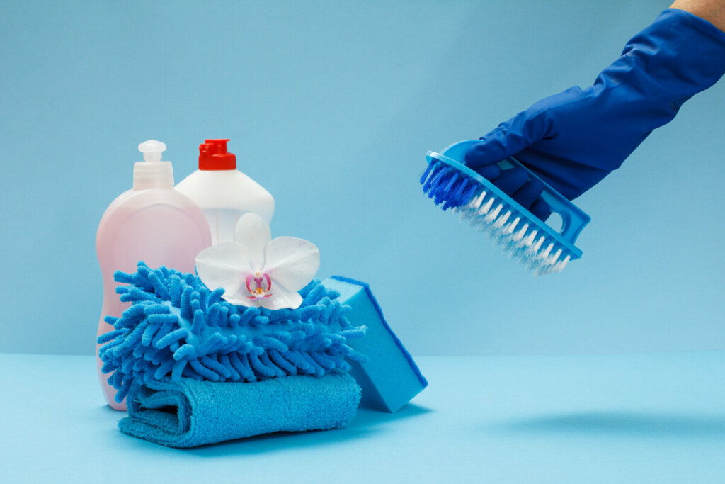Produtos de limpeza em cima de um fundo azul com uma mão com luva azul segurando uma escova de lavar roupa
