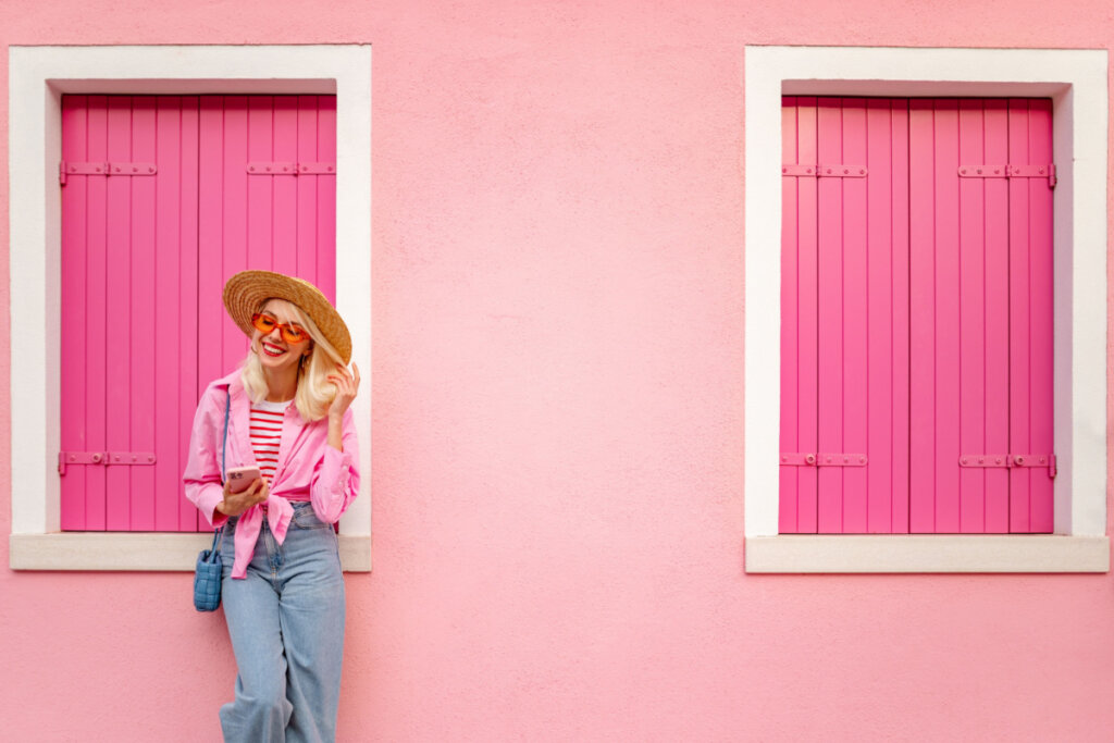 Viajante sorridente feliz usando chapéu elegante, óculos, camisa rosa, calças largas, andando, posando em uma parede cor de rosa