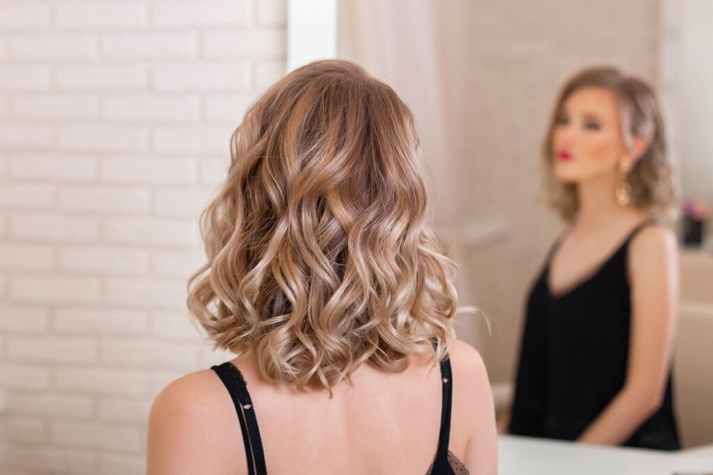 Mulher loira com cabelo ondulado de costas em frente a um espelho