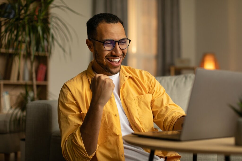 Homem com blusa branca e amarela sentado em frente a um computador sorrindo