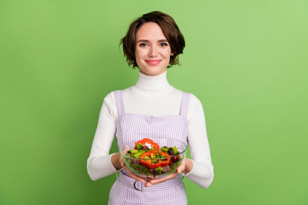 Mulher sorrindo segurando um recipiente com salada em frente a um fundo verde