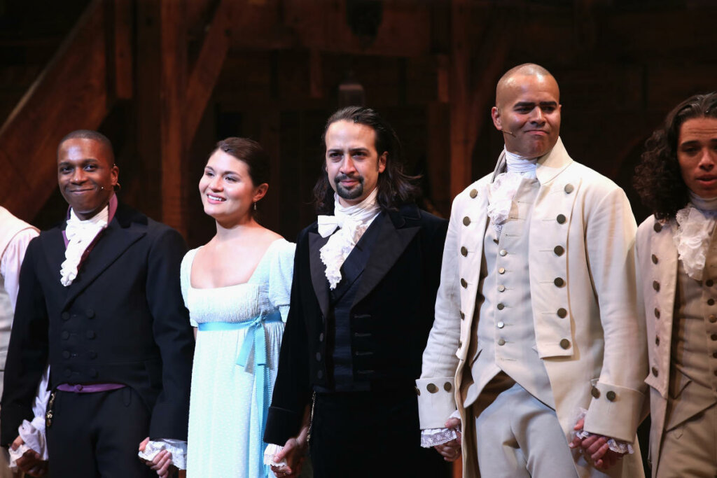 Cena do filme musical "Hamilton", com os personagens alinhados no palco sorrindo.