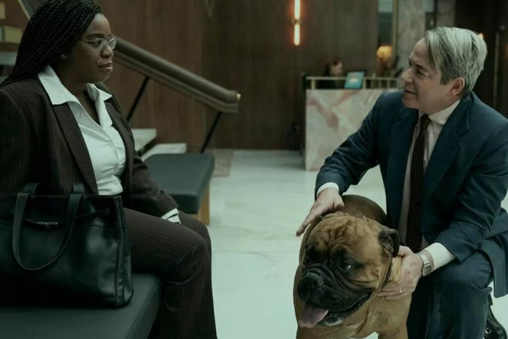 Cena da série "Império da Dor"; homem segurando cachorro e mulher negra sentada em sofá