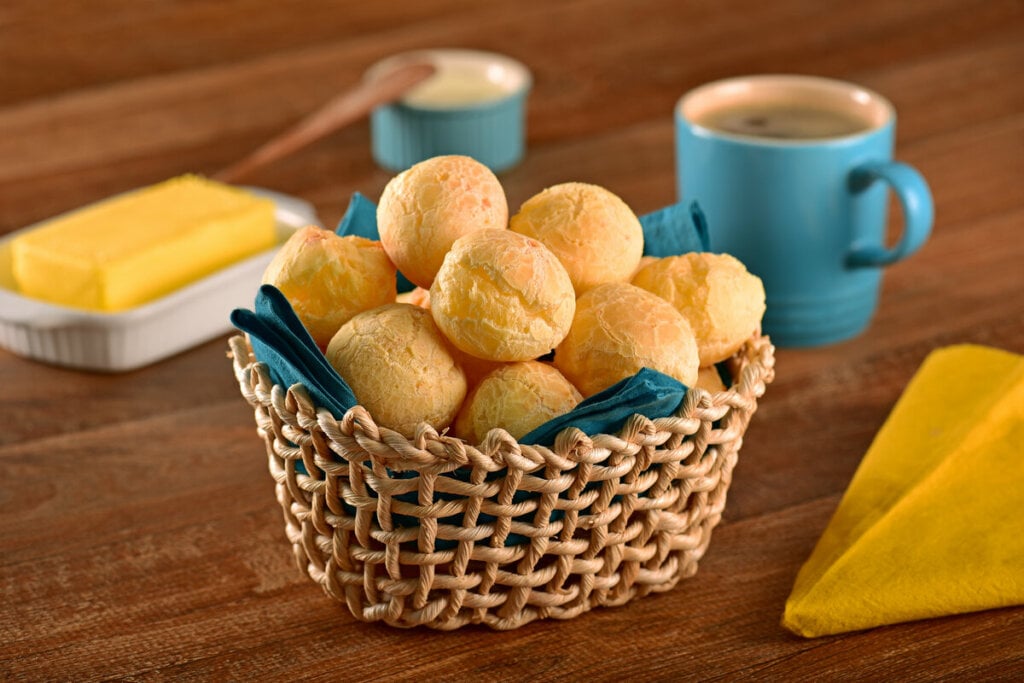 Cesta de palha forrada com pano verde com pães de queijo em cima de uma mesa de madeira