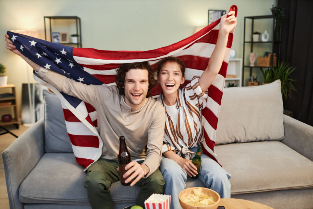 Retrato de um jovem casal com a bandeira dos EUA assistindo TV em casa, comendo e bebendo