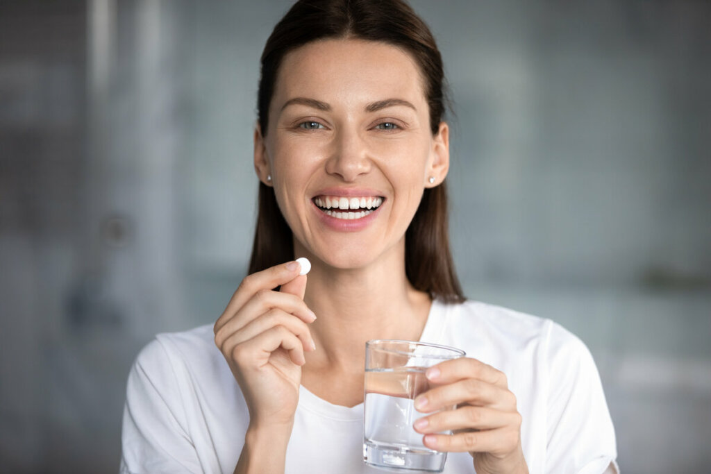 Mulher sorrindo segurando um copo com água e cápsula de remédio