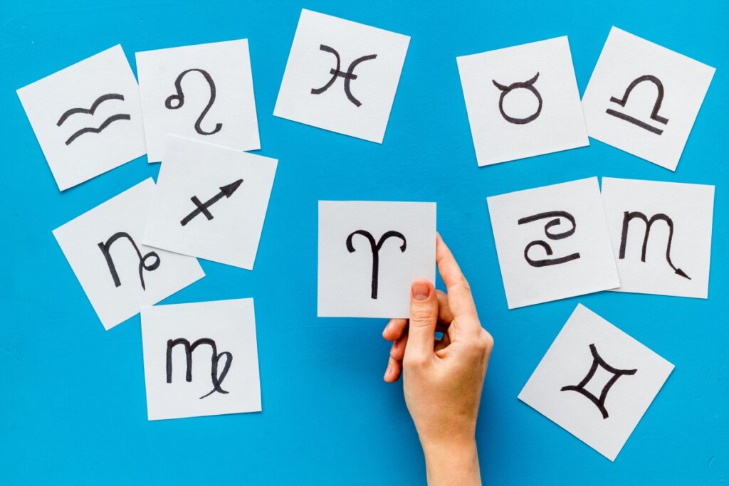 Mão feminina segurando um papel com o signo de Áries em um fundo azul com desenhos dos doze signos do zodíaco
