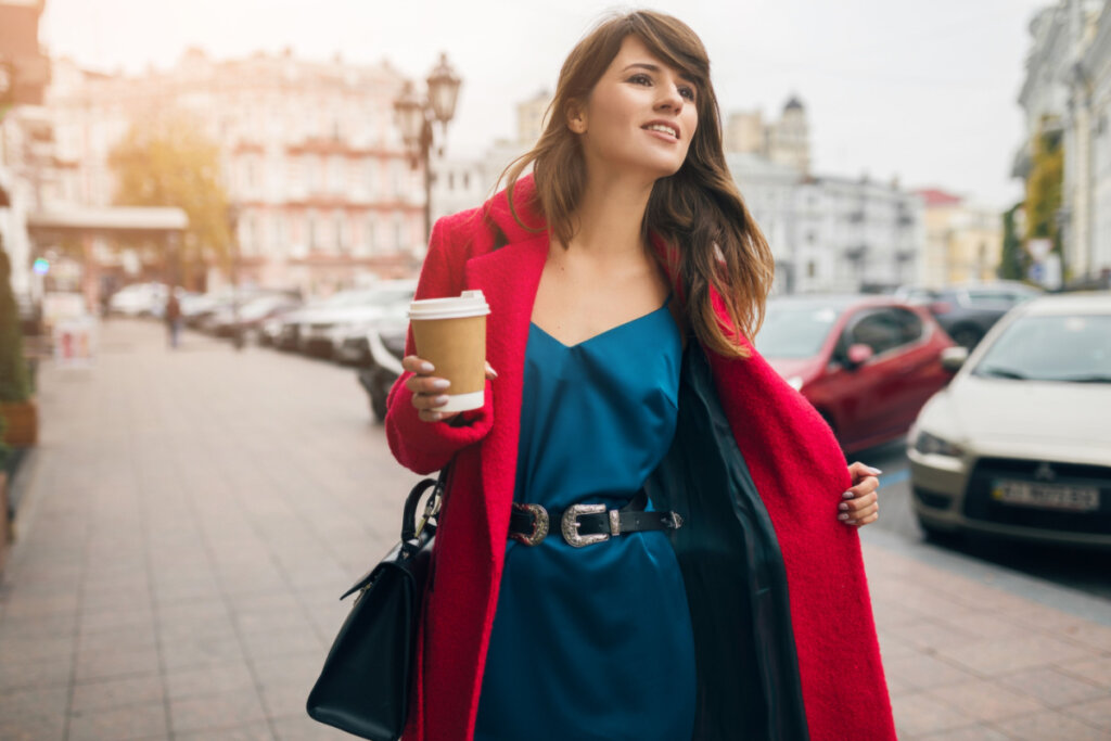 Mulher de vestido azul e casaco vermelho andando na rua com café na mão 
