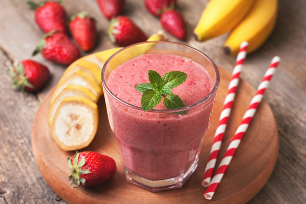 Copo com vitamina rosa decorado com folhas de hortelã em cima de uma tábua de madeira com morangos e rodelas de banana ao seu redor