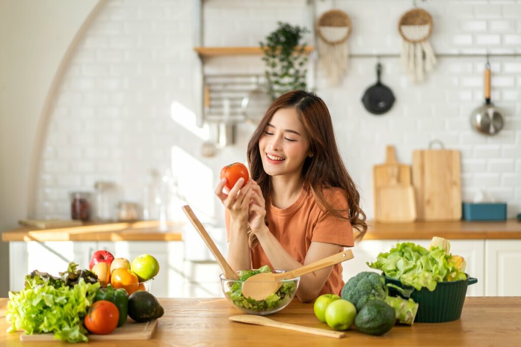 Mulher sorrindo escorada na bancada de uma cozinha segurando um tomate 