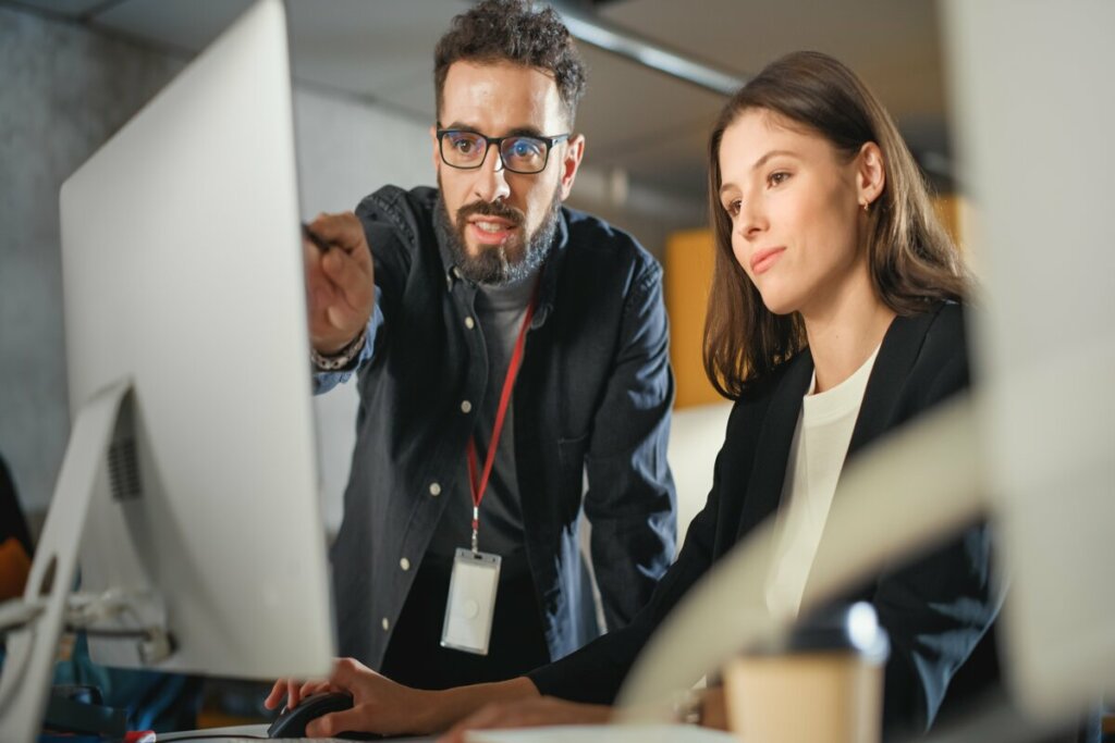 Mulher usando blazer preto sentada com um homem usando camisa social e óculos ao seu lado apontando para a tela de um computador