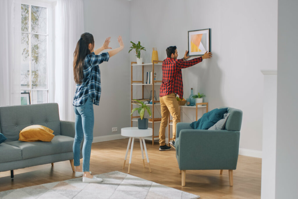 Homem e mulher pendurando um quadro na parede de uma sala de estar com poltrona verde e vasos de planta