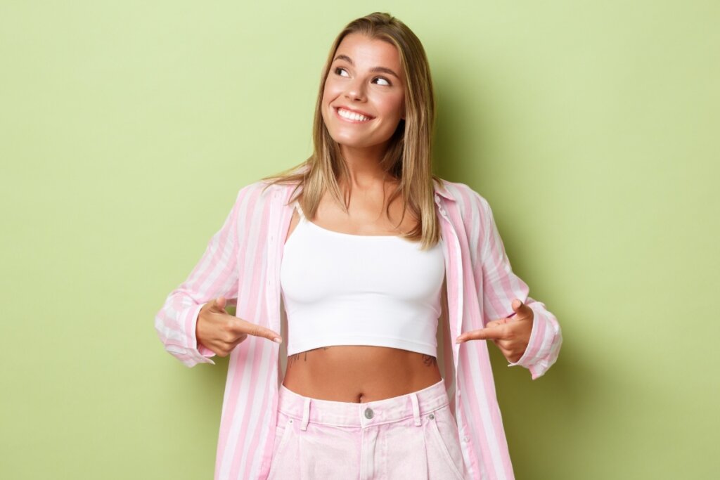 Mulher vestindo blusa branca , short e casaco rosa sorrindo e apontando para a barriga