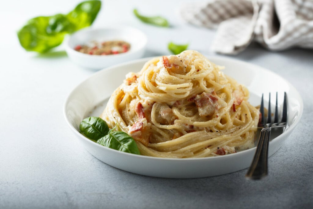 Prato branco com macarrão à carbonara em cima de uma mesa branca e decorado com folhas