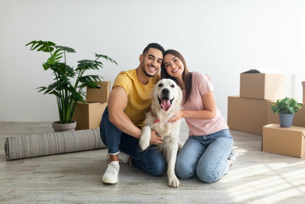 Homem e mulher abraçados com um cachorro em uma sala de estar com caixas de papelão