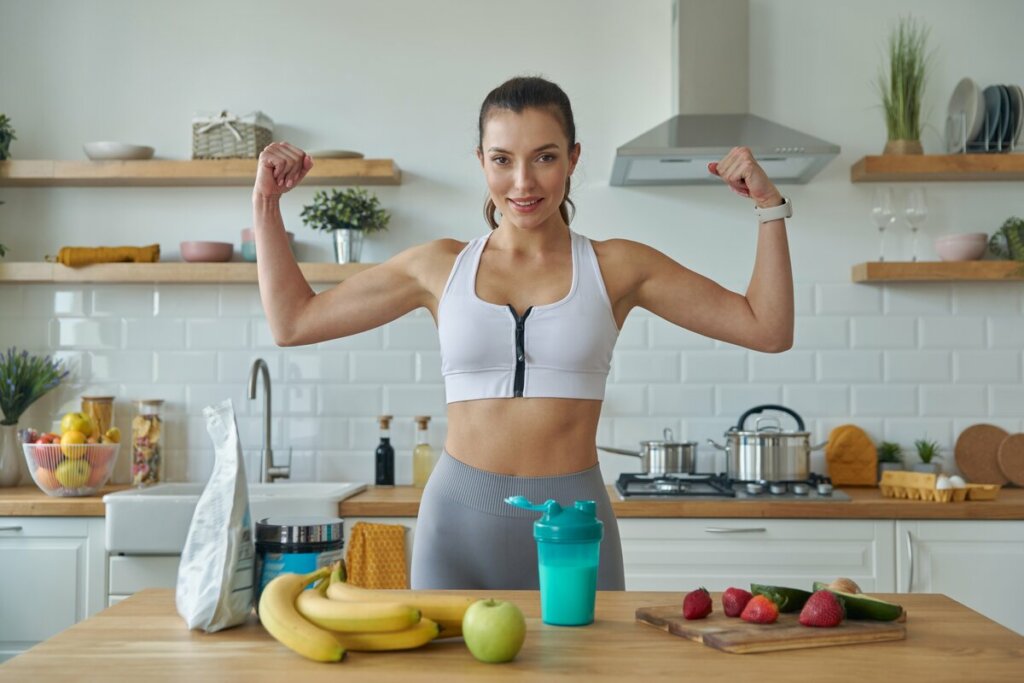 Mulher mostrando os músculos do braço em uma cozinha com mesa com frutas e whey protein