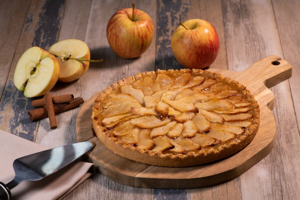Torta de maçã em cima de tábua de madeira com maçãs e canelas em paus ao lado
