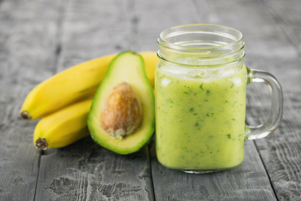 Copo de vidro com vitamina de abacate em cima de uma mesa de madeira com duas bananas e um abacate