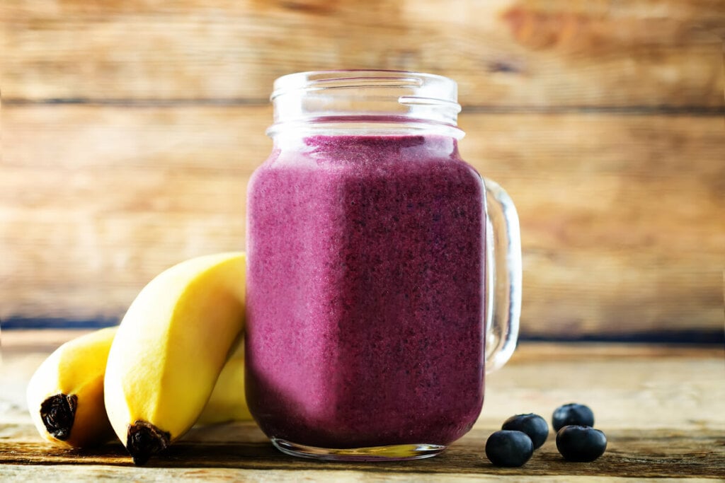 Copo de vidro com vitamina de açaí e banana em cima de uma mesa de madeira com banana e açaí ao lado