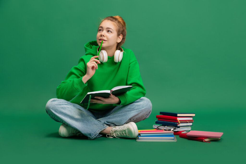 Estudante sentada no chão com expressão pensativa, estudando isolada sobre fundo verde do estúdio