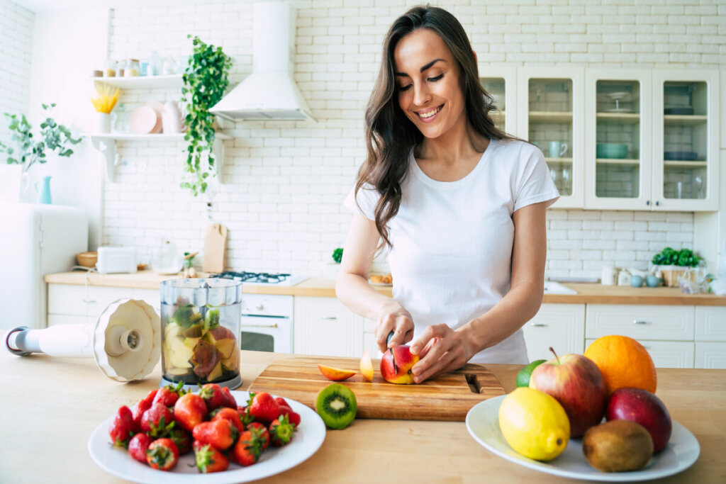 Mulher vestindo blusa branca em pé em uma cozinha, cortando frutas e sorrindo 