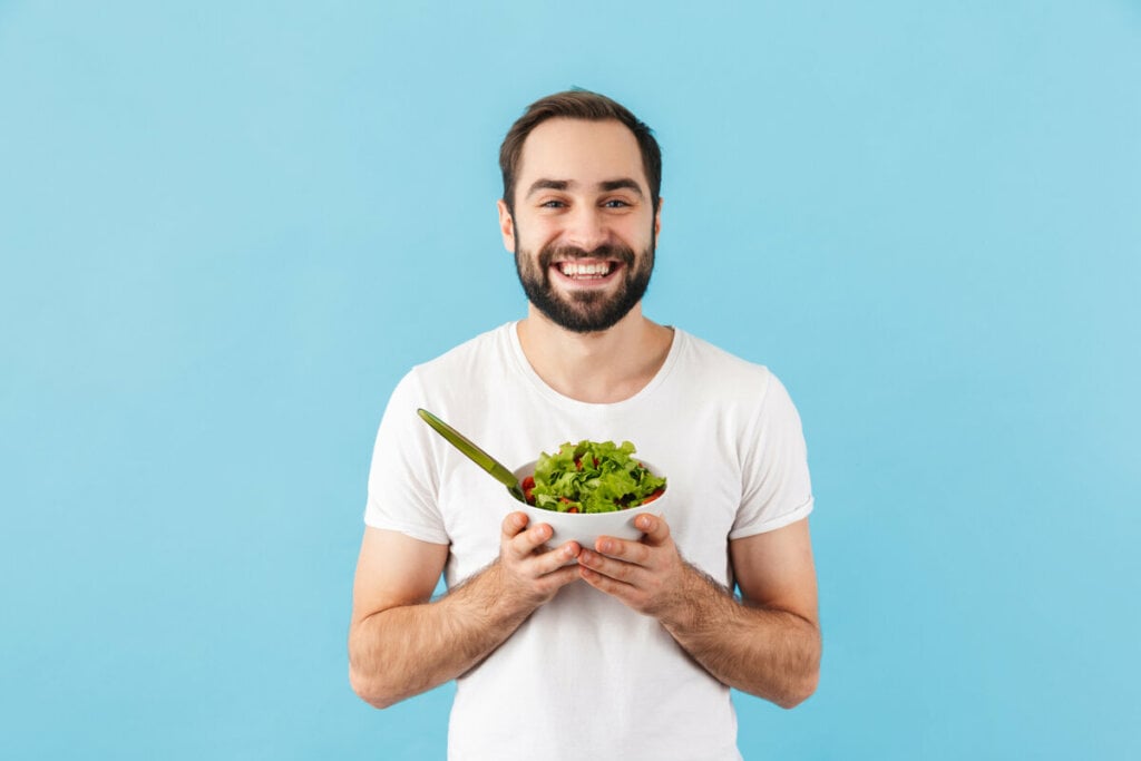 Homem vestindo blusa branca em frente a um fundo azul, sorrindo e segurando um recipiente com salada