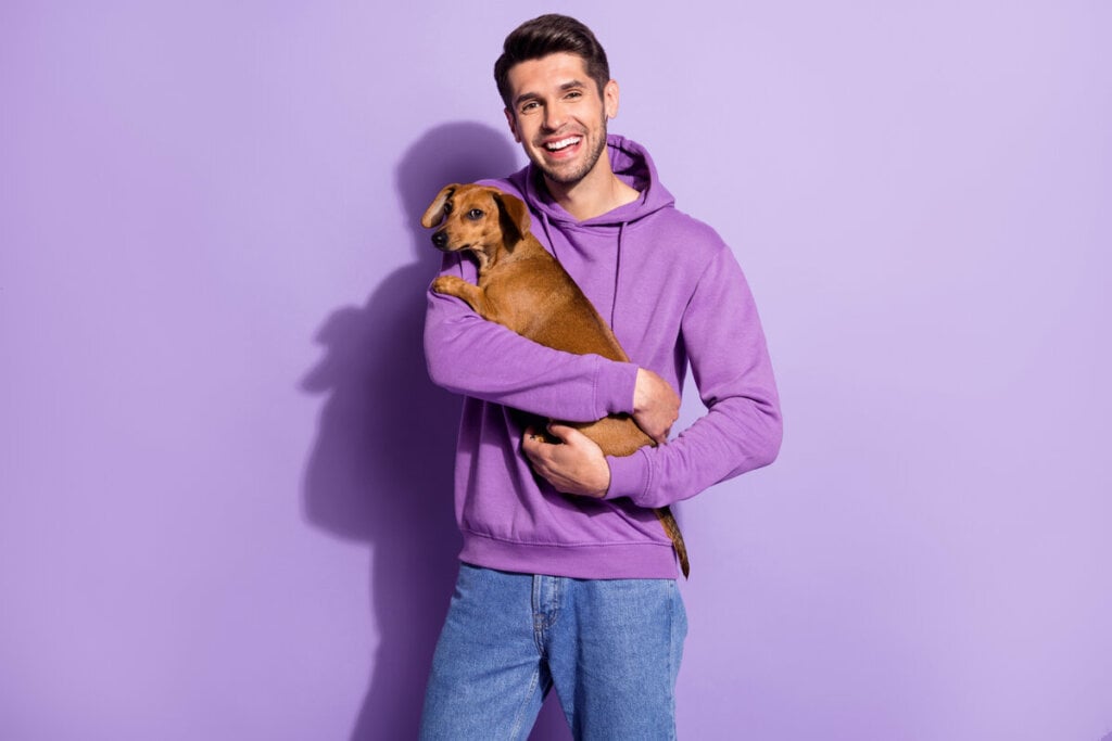 Homem vestindo blusa de moletom roxa e segurando um cachorro em frente a um fundo roxo