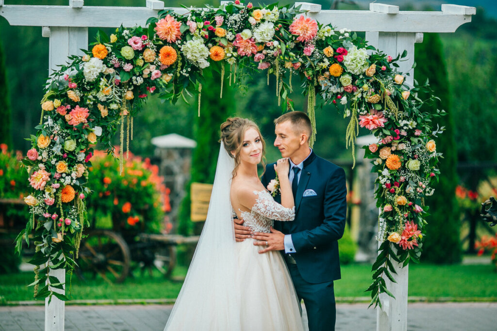 Mulher vestida de noiva e homem com terno em frente a um arco de flores abraçados