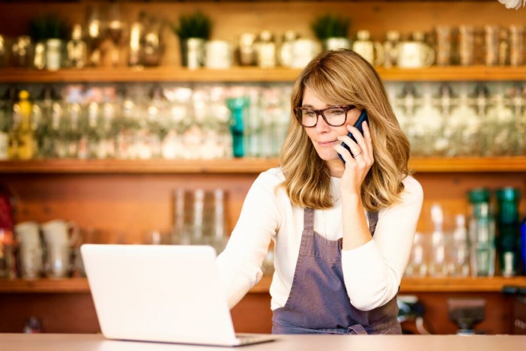 Mulher vestindo avental sentada mexendo em um computador e falando no telefone
