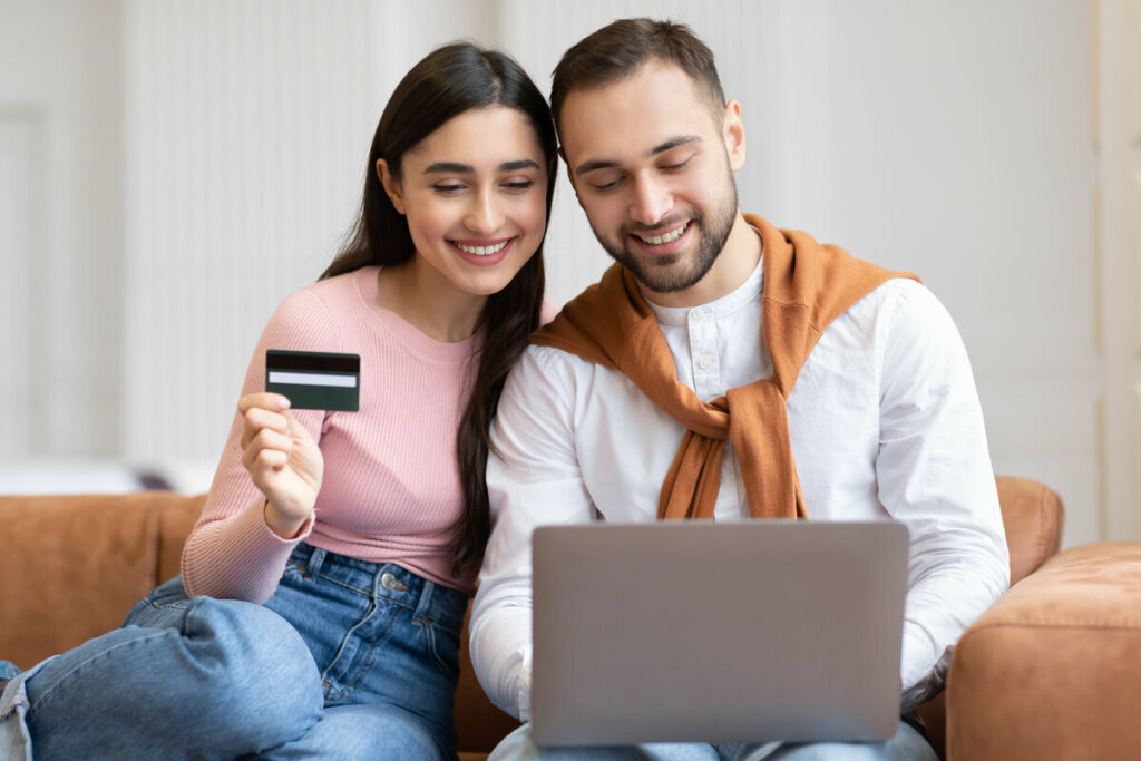 Homem e mulher sentados olhando para a tela de um notebook com a mulher segurando um cartão de crédito 
