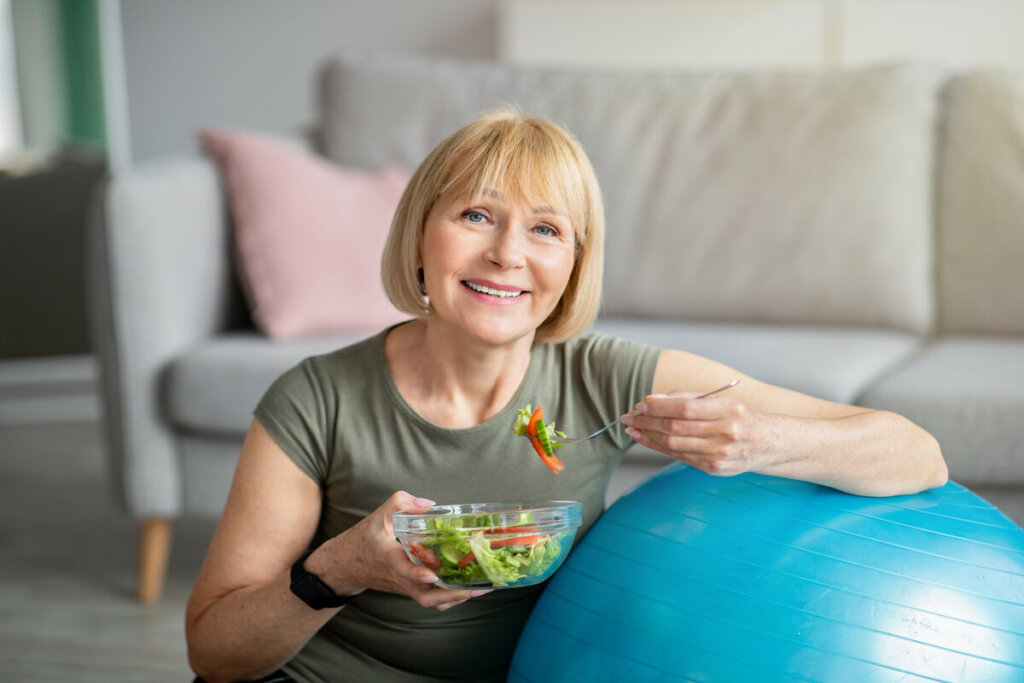 Mulher sentada com o braço em cima de uma bola azul e segurando um recipiente com salada 