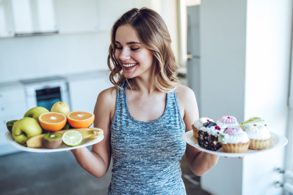 Mulher esportiva está escolhendo entre frutas e doces enquanto está em pé na cozinha luz.