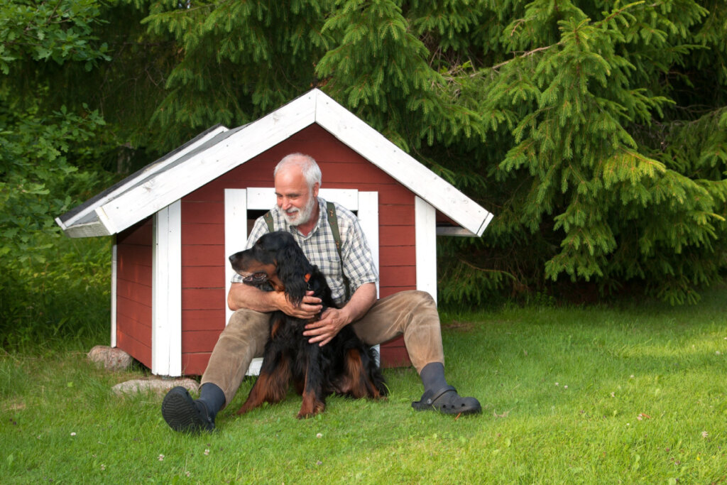 Homem sentado com cachorro preto em frente a casinha vermelha e branca