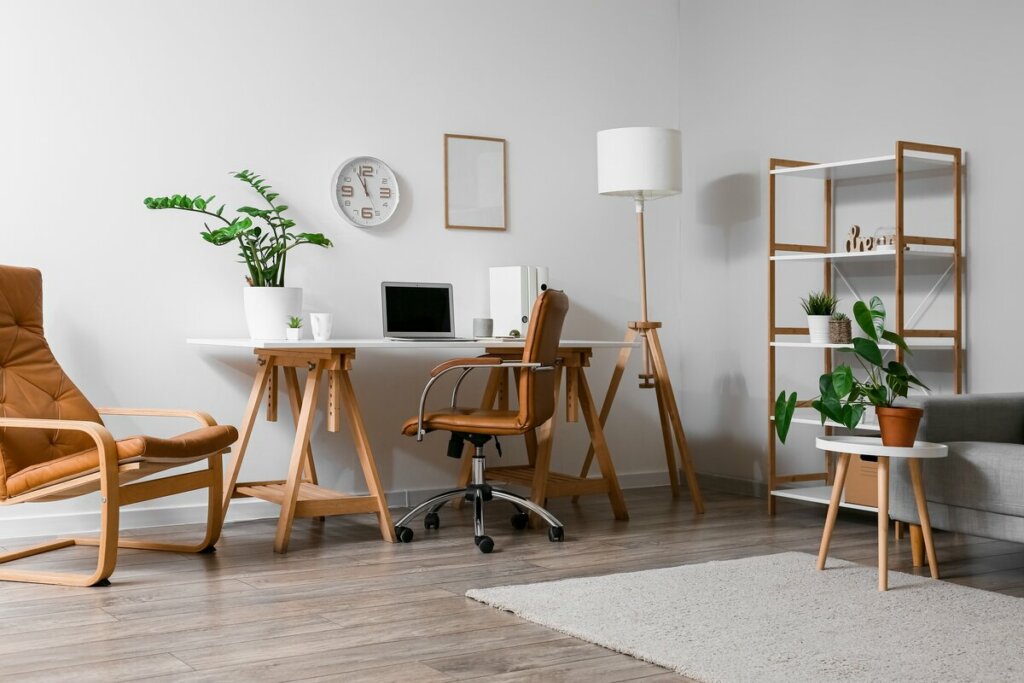 Sala de estar com parede branca, mesa de madeira com notebook, vasos de planta e uma cadeira marrom