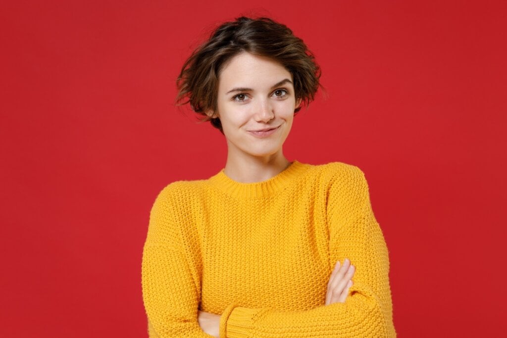 Mulher vestindo com blusa amarela com os braços cruzados em frente a um fundo vermelho